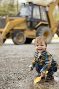 A young boy plays in a puddle with a shovel in front of an excavator.