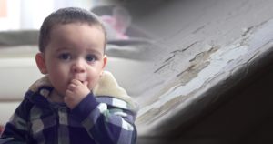 Child touches his mouth and sits in front of structure with chipping paint. 
