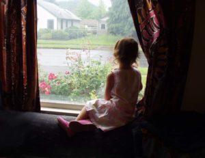 Young girl sits by the window and looks out into the yard.